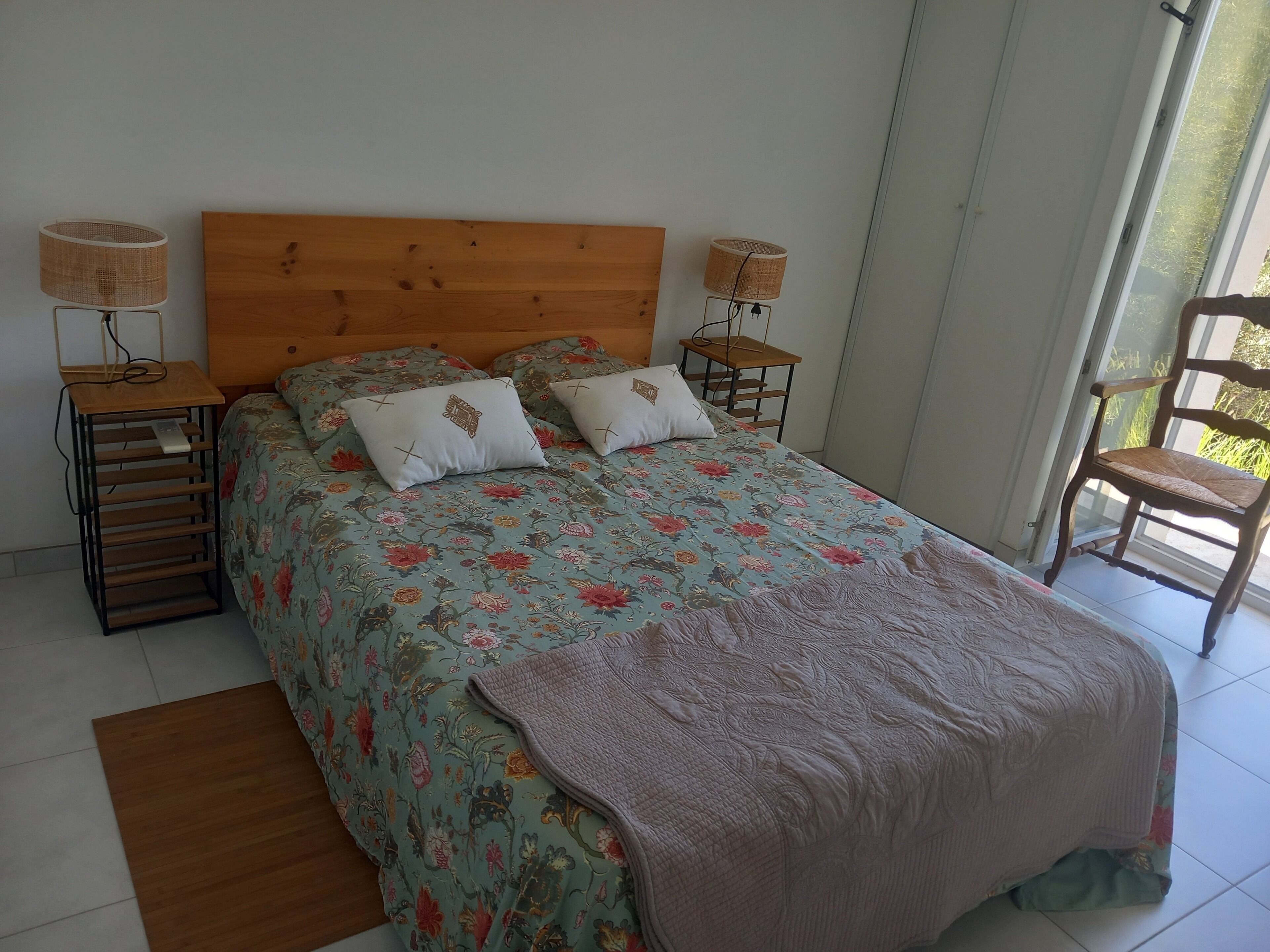 A bedroom with a wooden headboard, floral bedding, two bedside lamps, a wooden nightstand, a chair, and a window with a view.