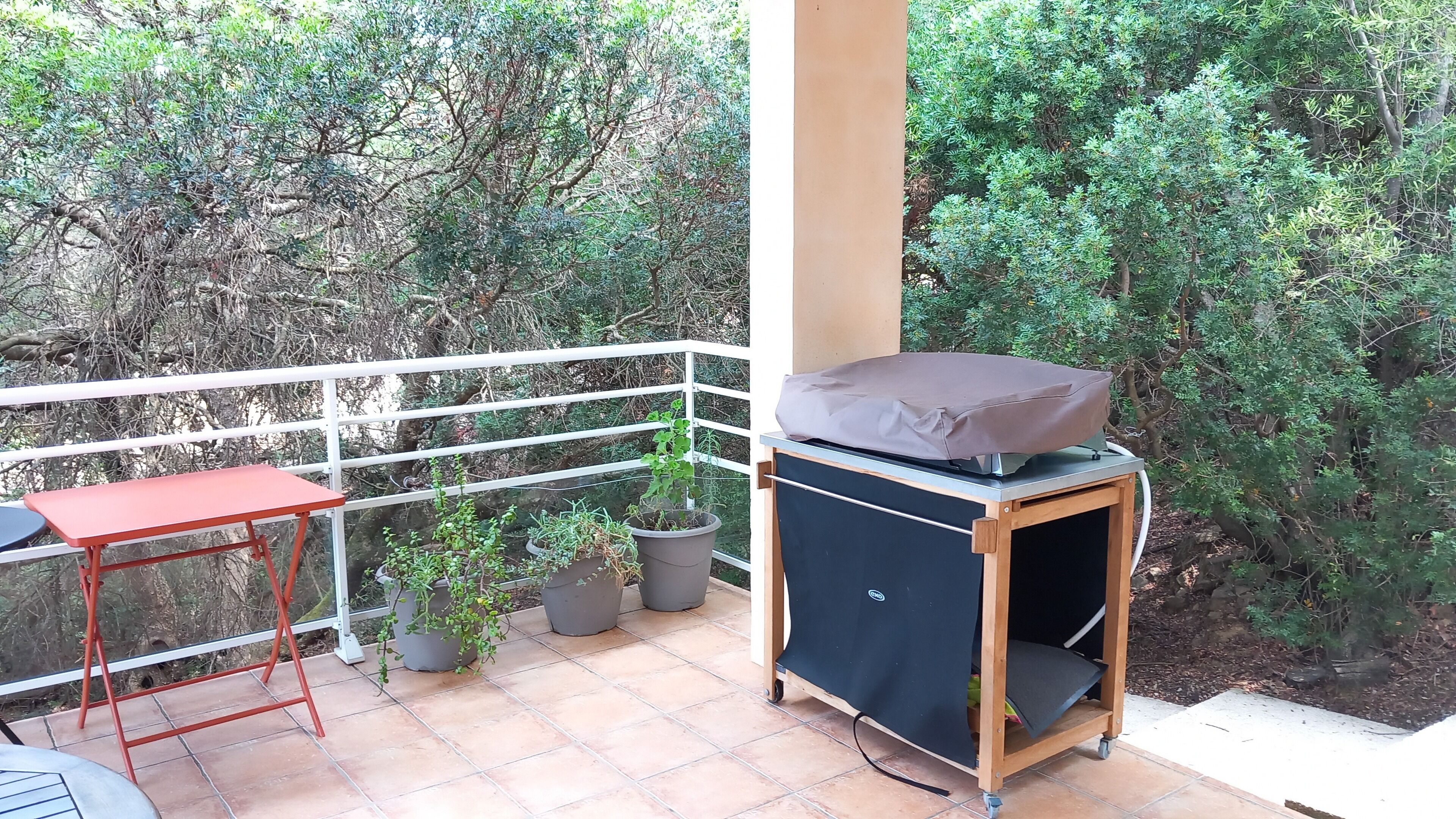A balcony with a red table, potted plants, and a covered grill.