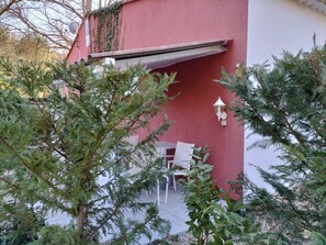 Un edificio rojo con una silla y una mesa blancas afuera, rodeado de árboles perennes.