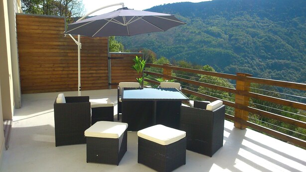 Een terras buiten met een tafel en stoelen, een houten hek en uitzicht op een bebost gebergte.