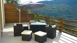 Een terras buiten met een tafel en stoelen, een houten hek en uitzicht op een bebost gebergte.