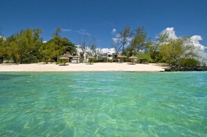 Une propriété située en bord de mer, avec une eau turquoise cristalline, une plage de sable et plusieurs constructions aux toits de chaume.