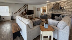 A spacious living room with a large grey sofa, a wooden coffee table, and a television on a white stand. A brick fireplace and a staircase are visible in the background.
