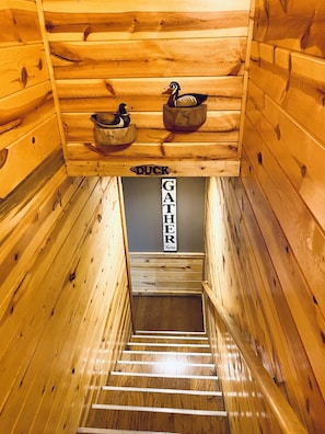 Ein Holzinnenraum mit einer Holztreppe, die zu einer unteren Ebene führt. Über der Treppe hängen zwei Holz-Eimer an der Wand, und ein Schild mit der Aufschrift 'DUCK GATHER HERE'.