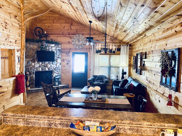 A cozy cabin interior with a stone fireplace, wooden walls, a dining area, and a living room furnished with a sofa and chairs.