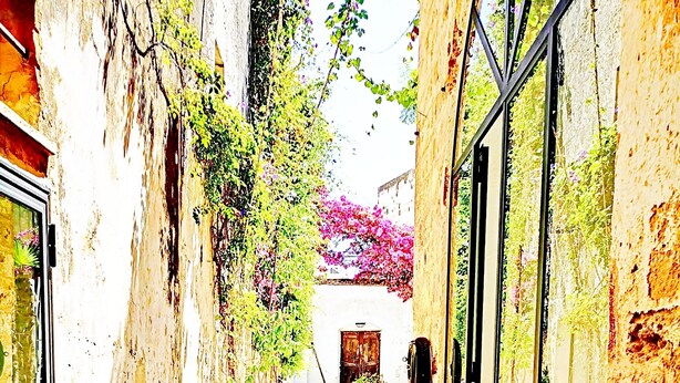 Une ruelle étroite aux murs de pierre, un tapis à motifs et une porte en bois au fond.