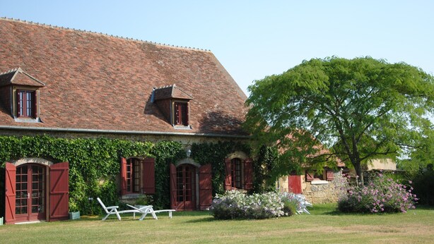 A traditional brick house with a sloped roof, a garden with white chairs, and a large tree.