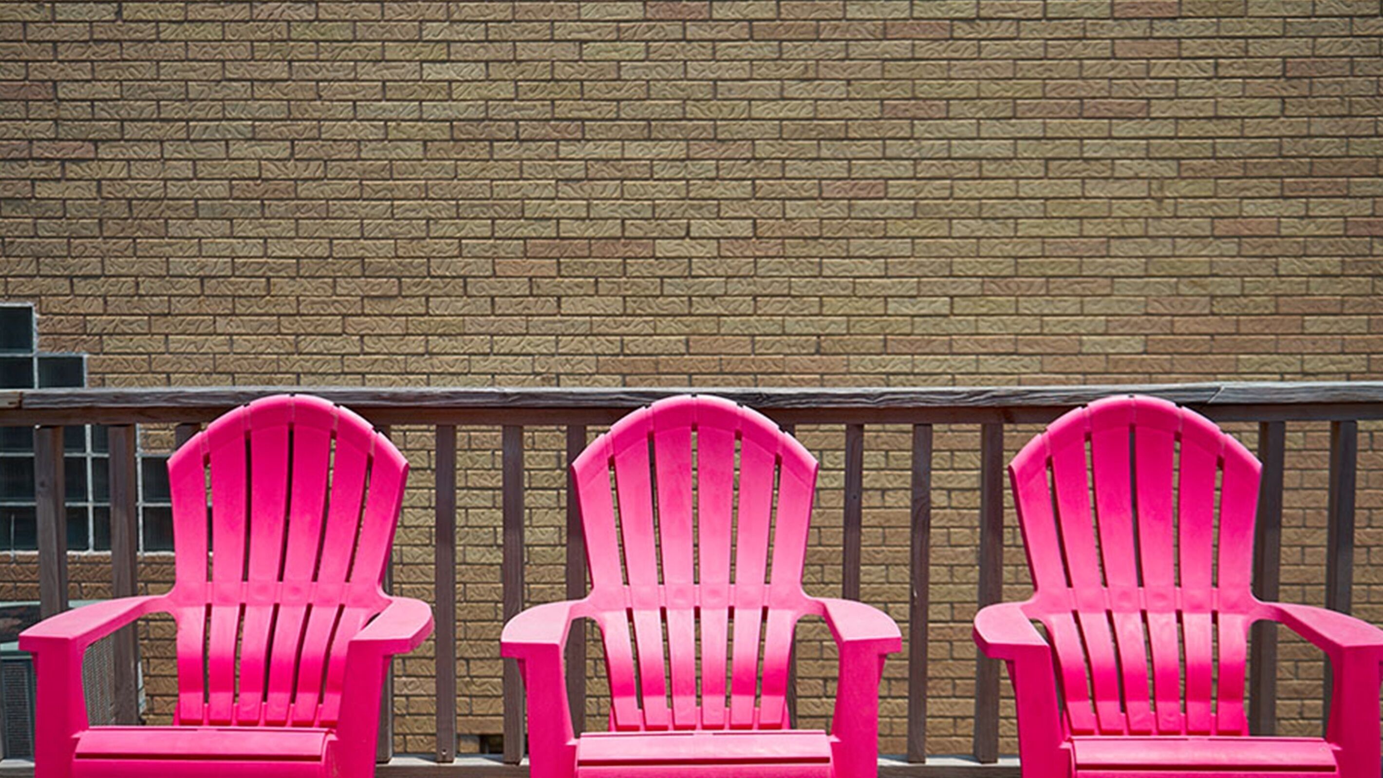 Three bright pink Adirondack chairs aligned against a brick wall.