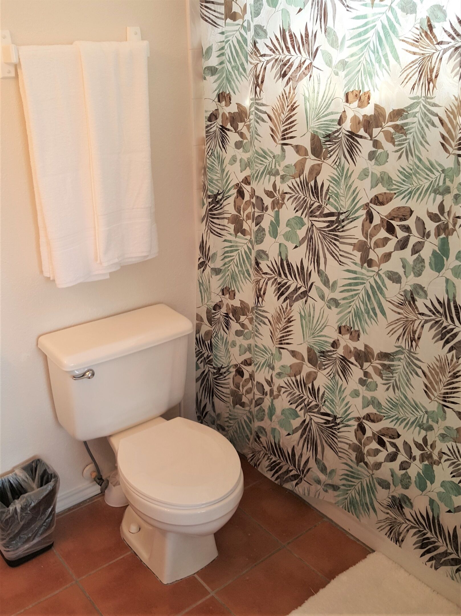 Een badkamer met een toilet, een prullenbak en een douchegordijn met een bladmotief.