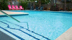 Una piscina con azulejos azules y un trampolín. Se observan sillas rosadas y una valla de madera al fondo.