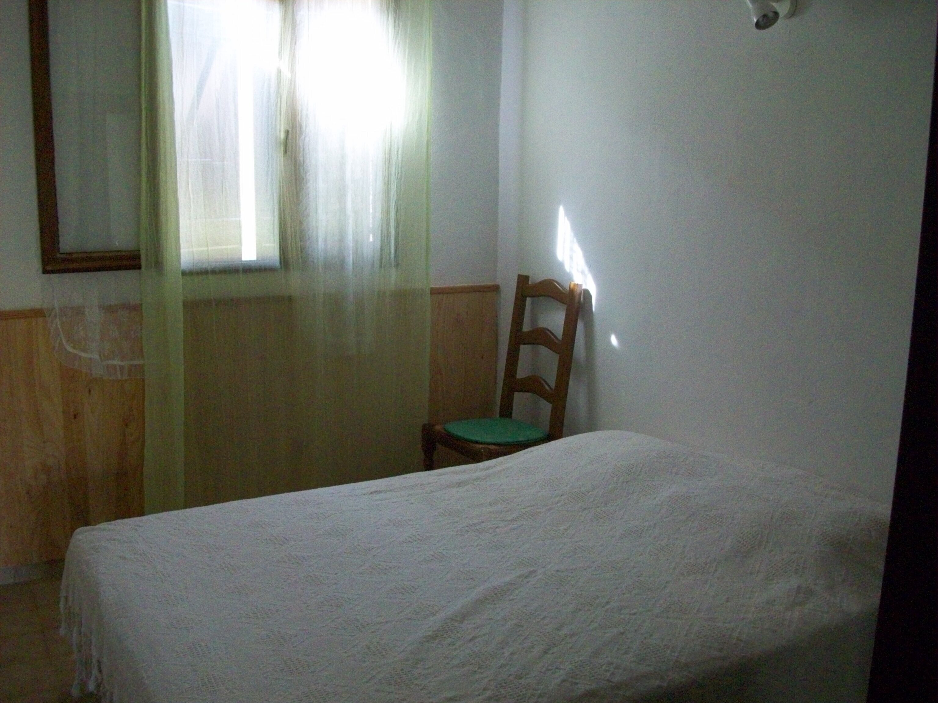 A small, simple room with a single bed, a wooden chair, and a window with sheer curtains.
