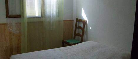 A small, simple room with a single bed, a wooden chair, and a window with sheer curtains.