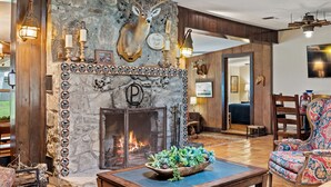 Une cheminée en pierre ornée d’une tête de cerf, avec un feu qui crépite et un décor ornemental. On y trouve une table en bois sur laquelle est posée une plante en guise de centre de table, ainsi qu’un fauteuil à motifs.