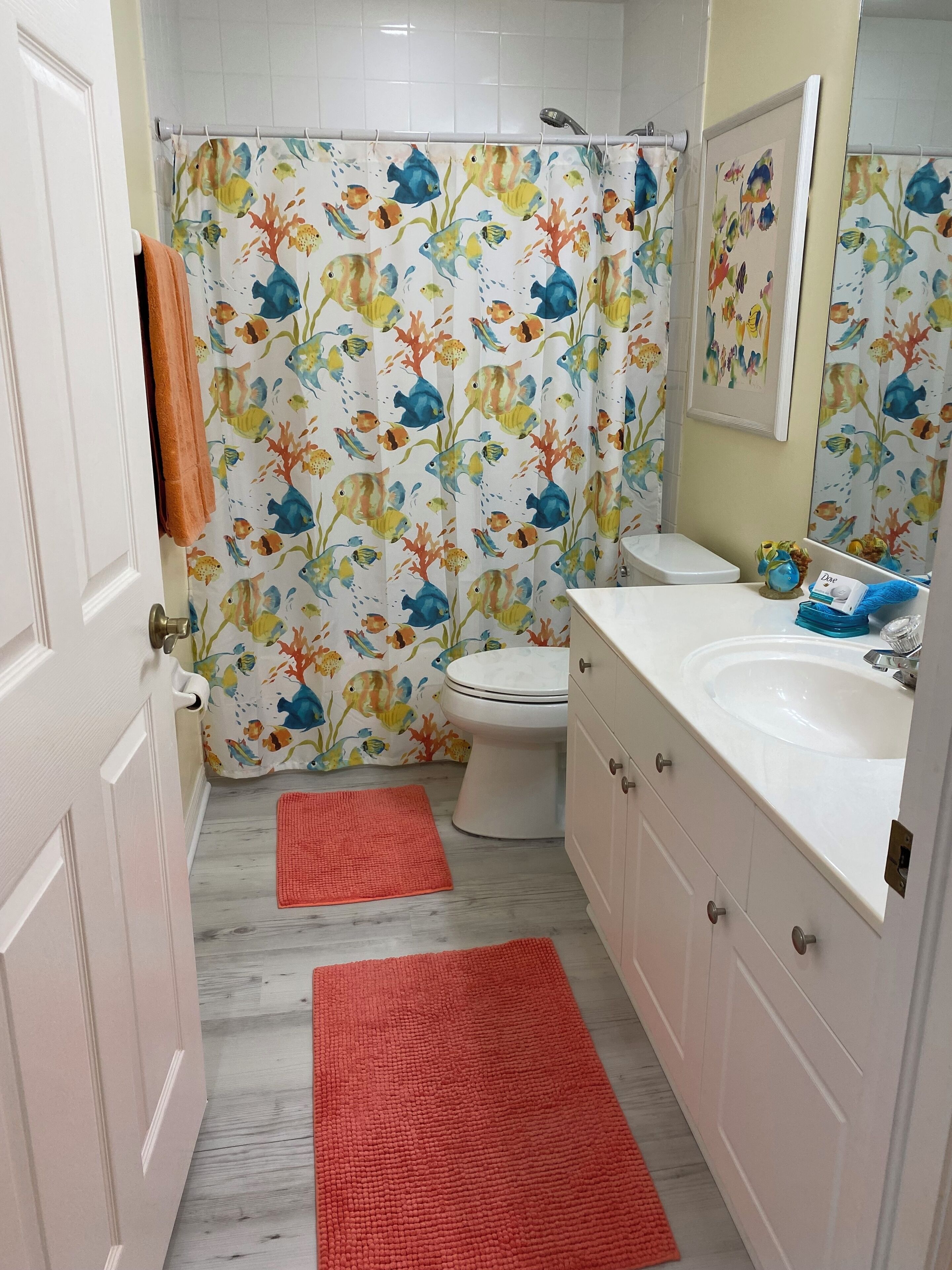 Un baño con cortina de ducha estampada con motivos de peces, un inodoro, un lavamanos y dos alfombras rojas en el suelo.
