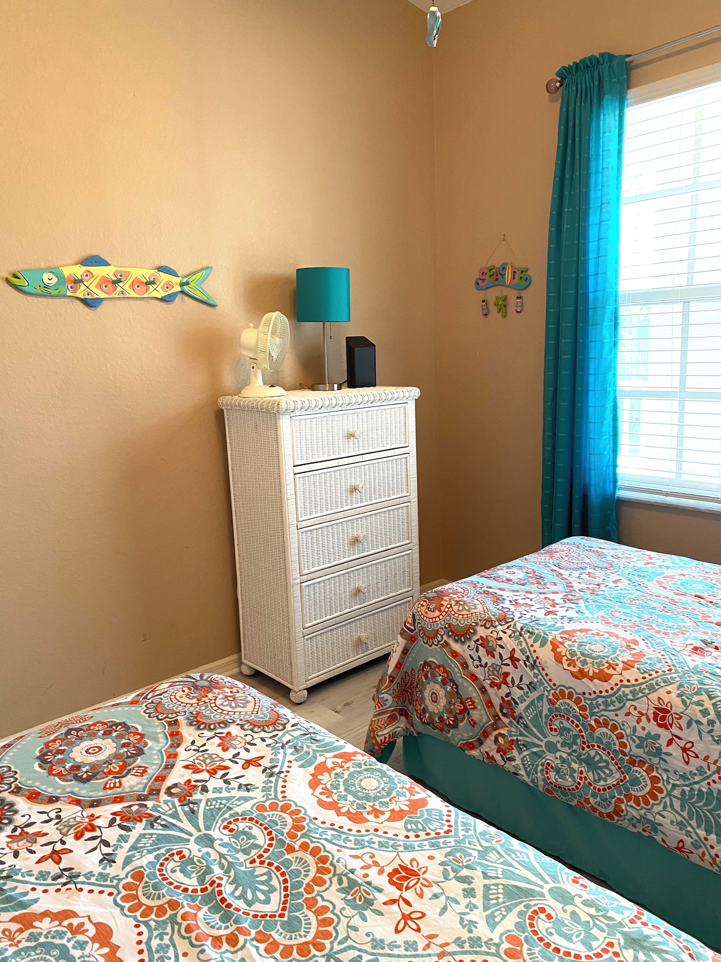 A bedroom with two beds, a dresser, a fan, a lamp, and a window with curtains.