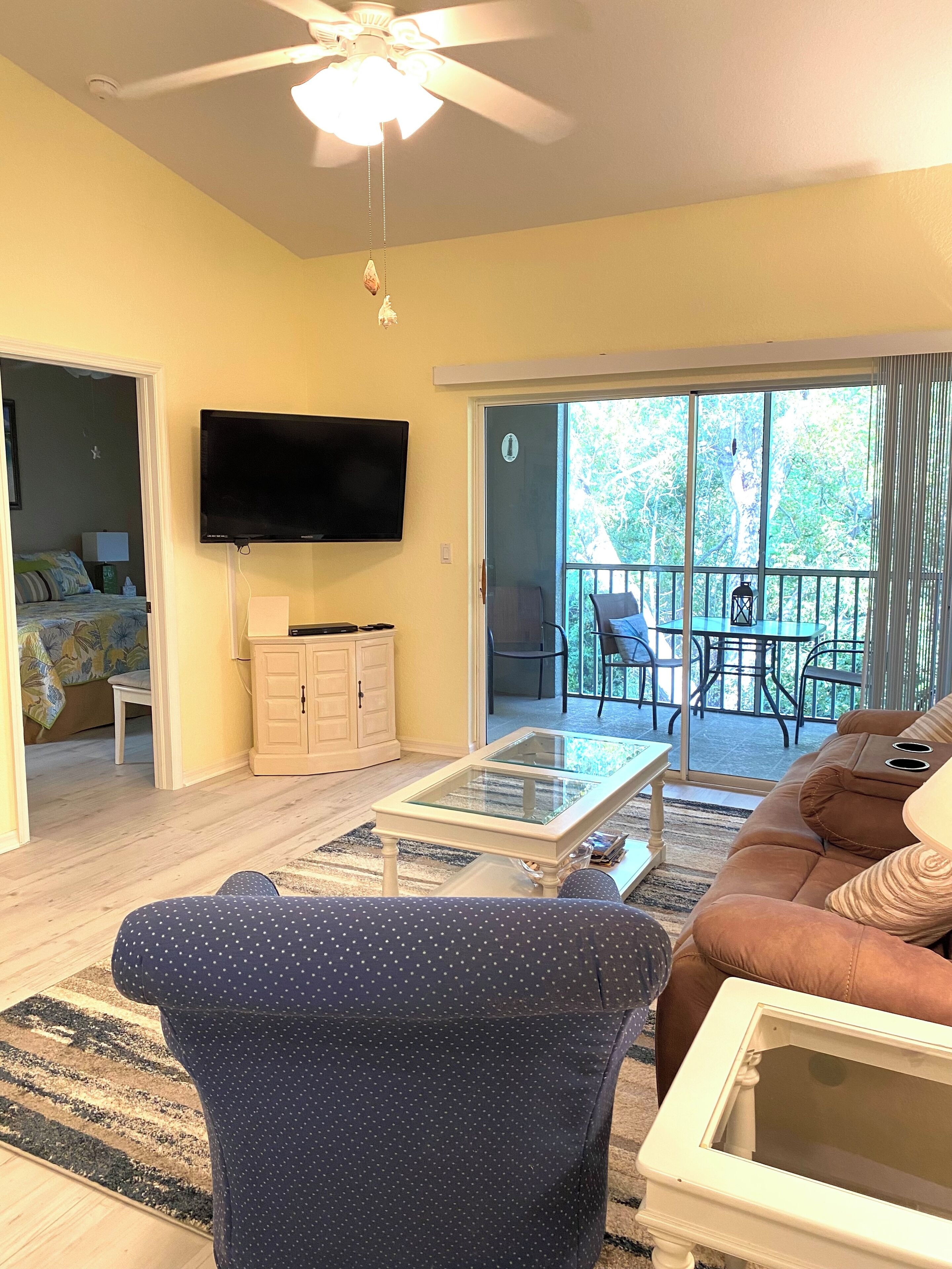 A living room with a ceiling fan, a flat-screen TV, a glass coffee table, a brown sofa, and a sliding glass door leading to a balcony with outdoor furniture.