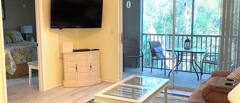 A living room with a ceiling fan, a flat-screen TV, a glass coffee table, a brown sofa, and a sliding glass door leading to a balcony with outdoor furniture.