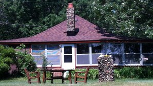 A house with a stone chimney, a red roof, and a large window. There is a wooden table and chairs in the front yard.