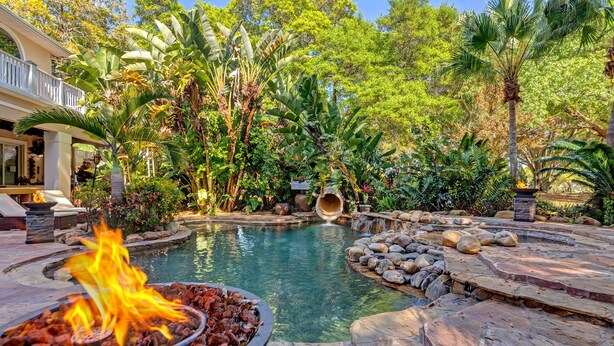 Zona de piscina con una chimenea escultural, rodeada de plantas tropicales y un suelo de piedra.