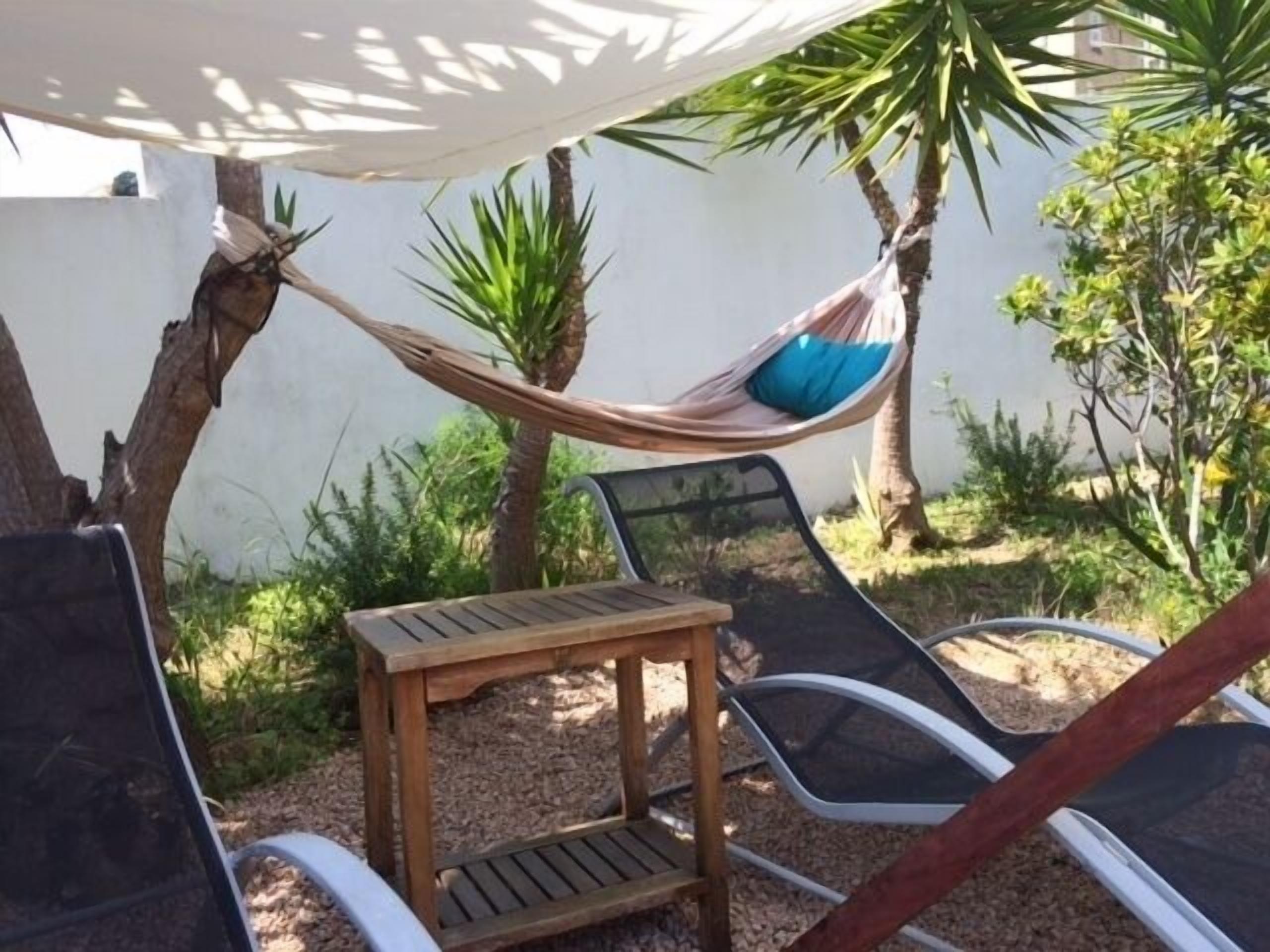 Eine Hängematte unter einer weißen Markise, zwei Liegestühle, ein kleiner Holztisch und ein Garten mit verschiedenen Pflanzen.