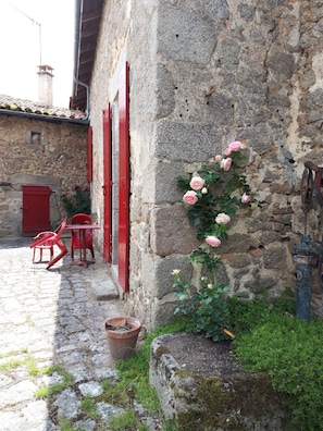Un bâtiment en pierre avec des volets rouges et un rosier. On y trouve des chaises rouges et une table à l’extérieur.