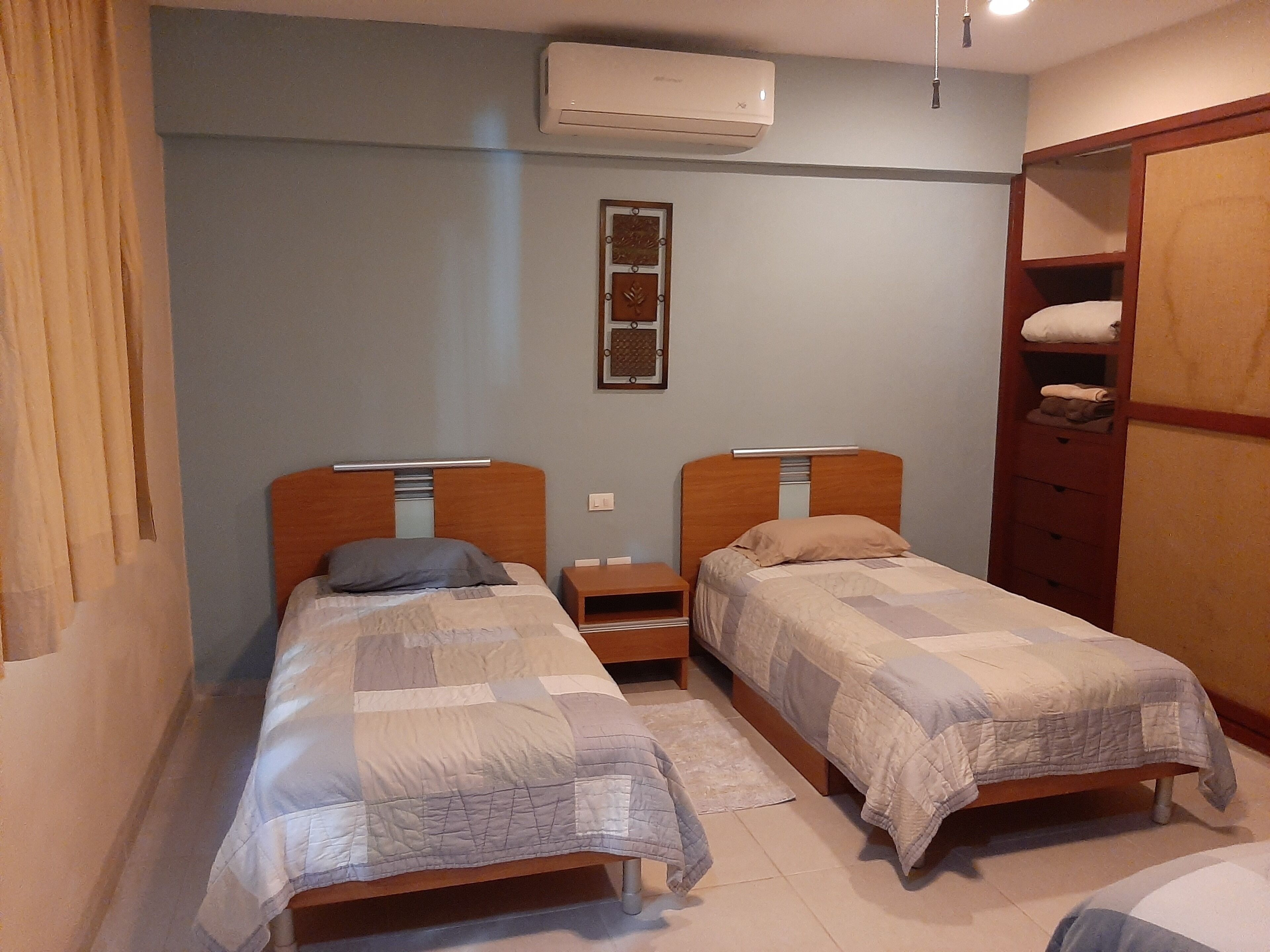 A bedroom with two single beds, a wooden wardrobe, a nightstand, and an air conditioning unit.