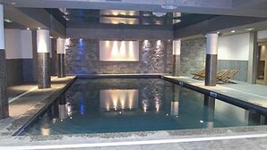 An indoor swimming pool with a stone wall backdrop, lounge chairs, and a tiled floor.