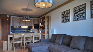 A cozy living room with a grey sofa, wooden dining table, and chairs. A kitchen with wooden cabinets and a hanging light fixture is visible in the background.
