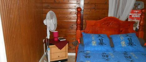 A wooden-paneled room with a bed, a standing fan, a small wooden chest, and a box labeled "VAPORMALL".