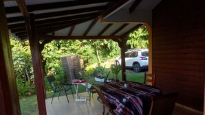 A covered patio with a table and chairs, surrounded by potted plants and a view of a parked car. - 2 min from the beach warm welcome in this flowered and wooded park (deshaies)