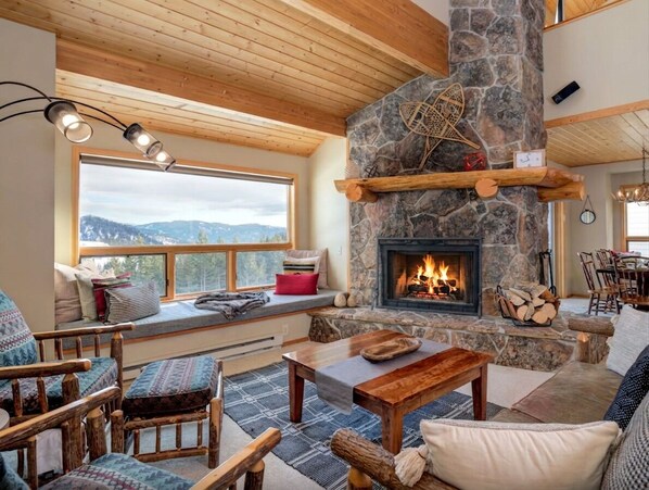 A cozy living room with a stone fireplace, wooden furniture, and a large window overlooking a mountainous landscape.