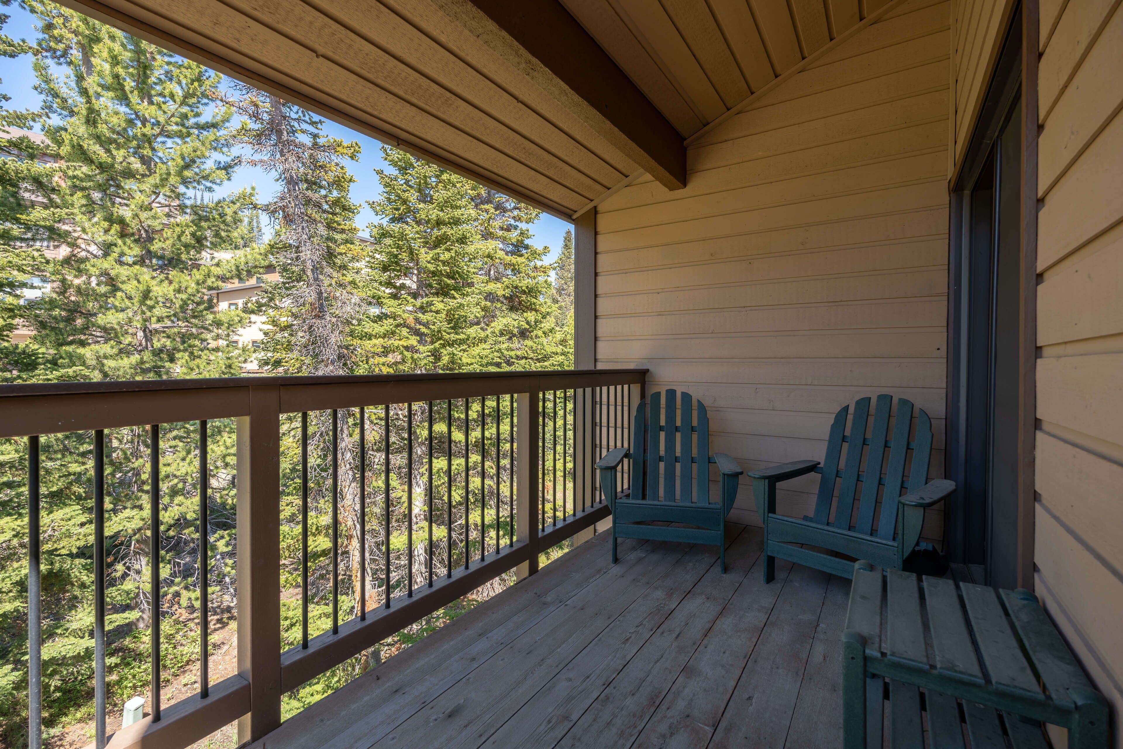 A wooden porch with two Adirondack chairs and a small table, overlooking a forested area.
