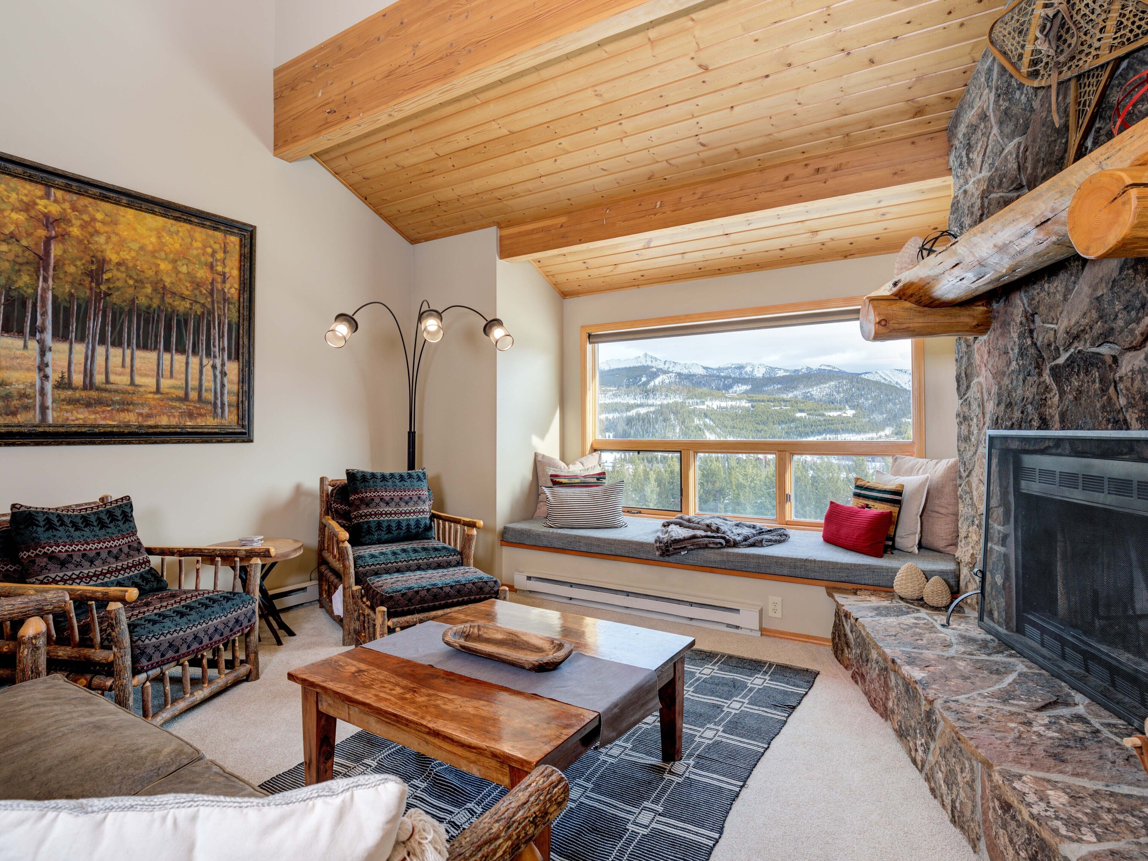 A cozy living room with a stone fireplace, wooden ceiling, and a large window offering a mountain view.