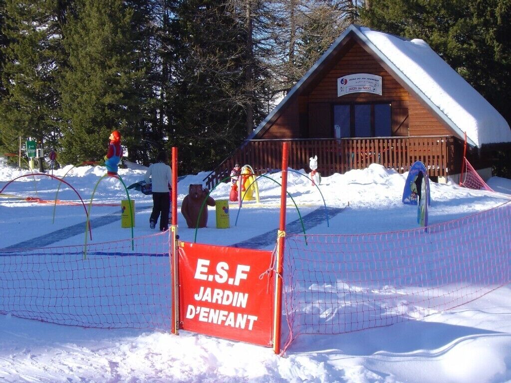 Une cabane en bois, dont le toit est recouvert de neige, entourée d’un espace délimité par une clôture et sur lequel est apposée une pancarte indiquant « E.S.F Jardin d’Enfant ».