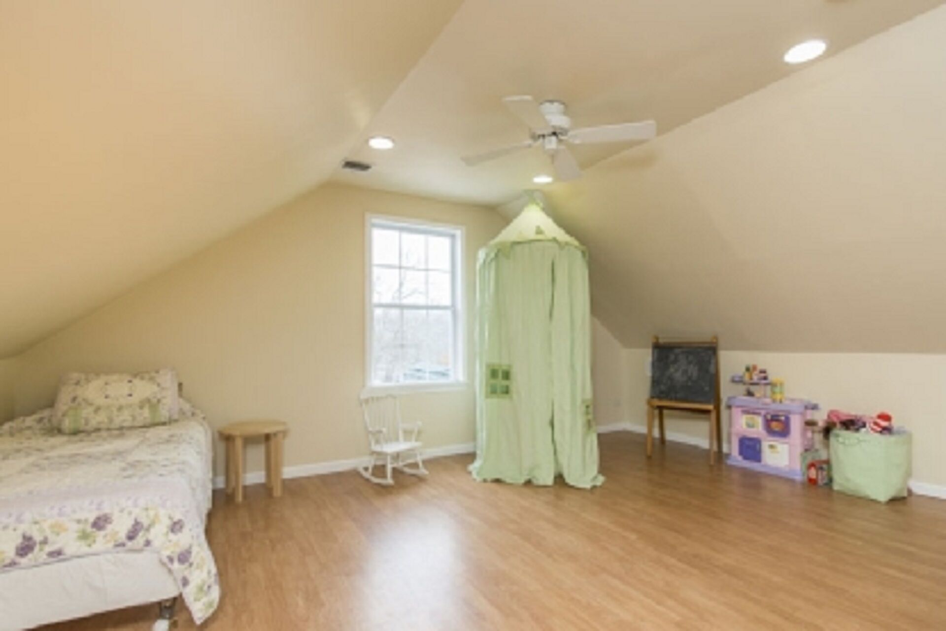 A bedroom with a sloped ceiling, a bed, a chair, a window, a ceiling fan, and a small table.