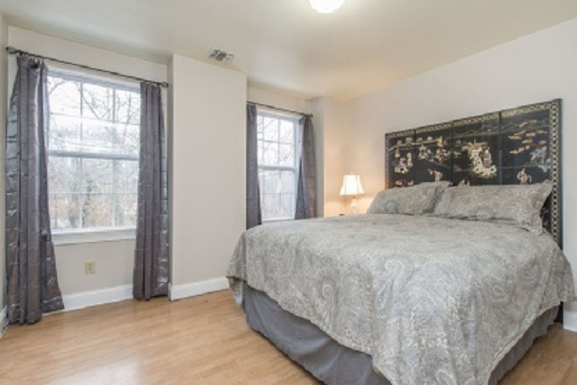 Une chambre à coucher avec un grand lit, deux fenêtres avec des rideaux, une lampe de chevet et une tête de lit agrémentée d’un panneau décoratif.