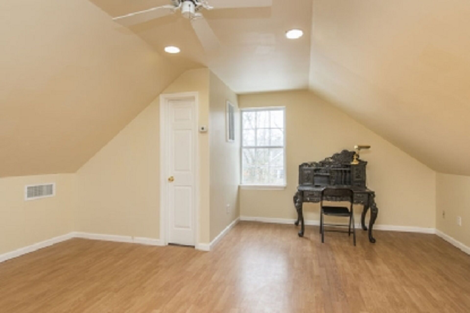 A spacious attic room with a sloped ceiling, a white door, a window, and a small table with a chair.