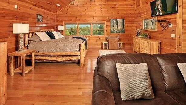 Interior de una cabaña de madera con una cama de madera, un sofá de cuero, un televisor y una chimenea.