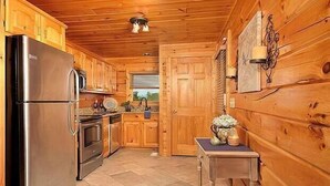 Cocina de cabaña de madera con refrigerador de acero inoxidable, armarios de madera, una pequeña mesa con un florero y una ventana con persianas.