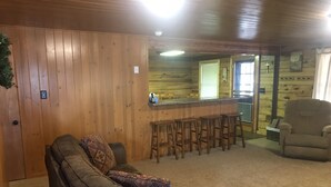 A cozy cabin interior with a leather sofa, a bar area with stools, and a wooden wall. - Family Friendly Lakefront Cedar Log Cabin on 2 acres at Rainbow Lake. (Lakeside)