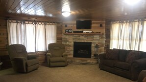 A cozy living room with a stone fireplace, a flat-screen TV mounted above it, and a comfortable seating area with a sofa and two armchairs. - Family Friendly Lakefront Cedar Log Cabin on 2 acres at Rainbow Lake. (Lakeside)