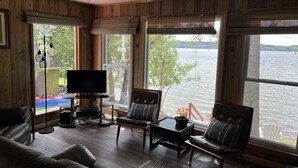 Une pièce chaleureuse aux murs en bois, avec une grande fenêtre donnant sur un lac, une télévision et des sièges confortables.