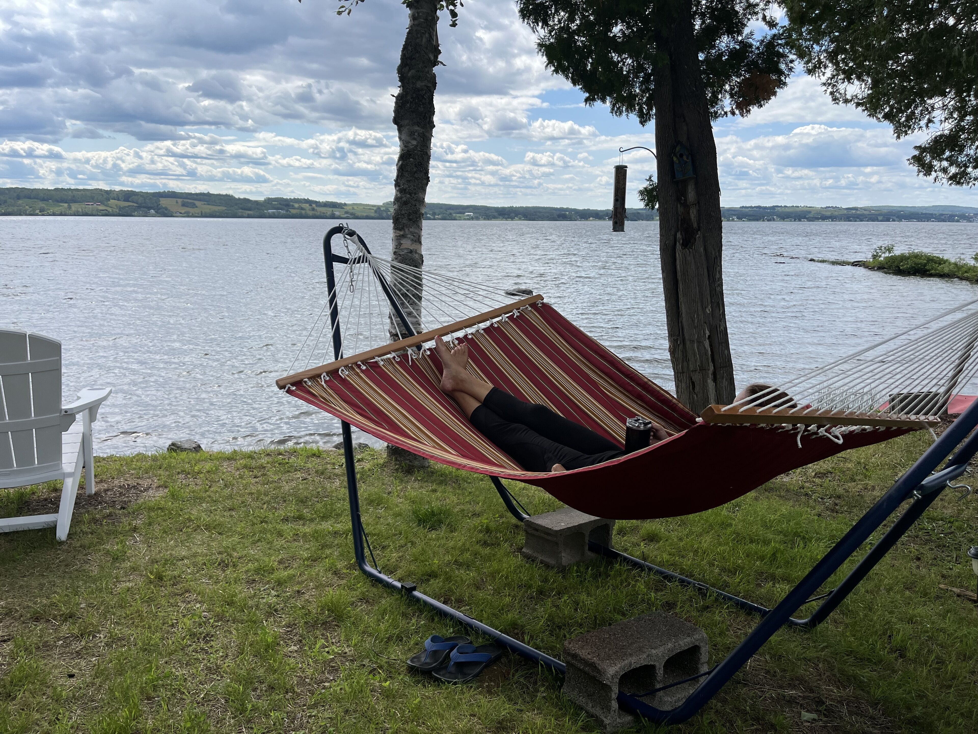A hammock with a person lying in it, a white chair, and a view of a lake with trees and a cloudy sky.