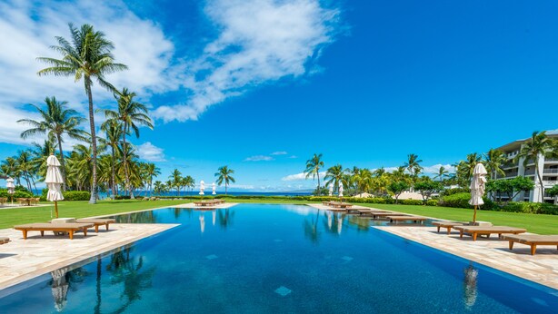A pool with lounge chairs and umbrellas, surrounded by palm trees and a clear blue sky.
