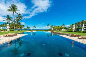 Piscina com espreguiçadeiras e guarda-sóis, cercada por palmeiras e um céu azul claro.