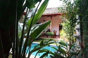 A pool area with a tiled roof structure, surrounded by lush greenery and potted plants.