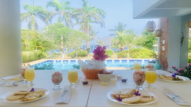 A table set with breakfast items including toast, fruit, and juice, overlooking a pool and palm trees.