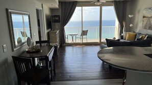 Uma sala de estar moderna com uma janela grande voltada para o oceano, um ventilador de teto e uma área de jantar com mesa e cadeiras.