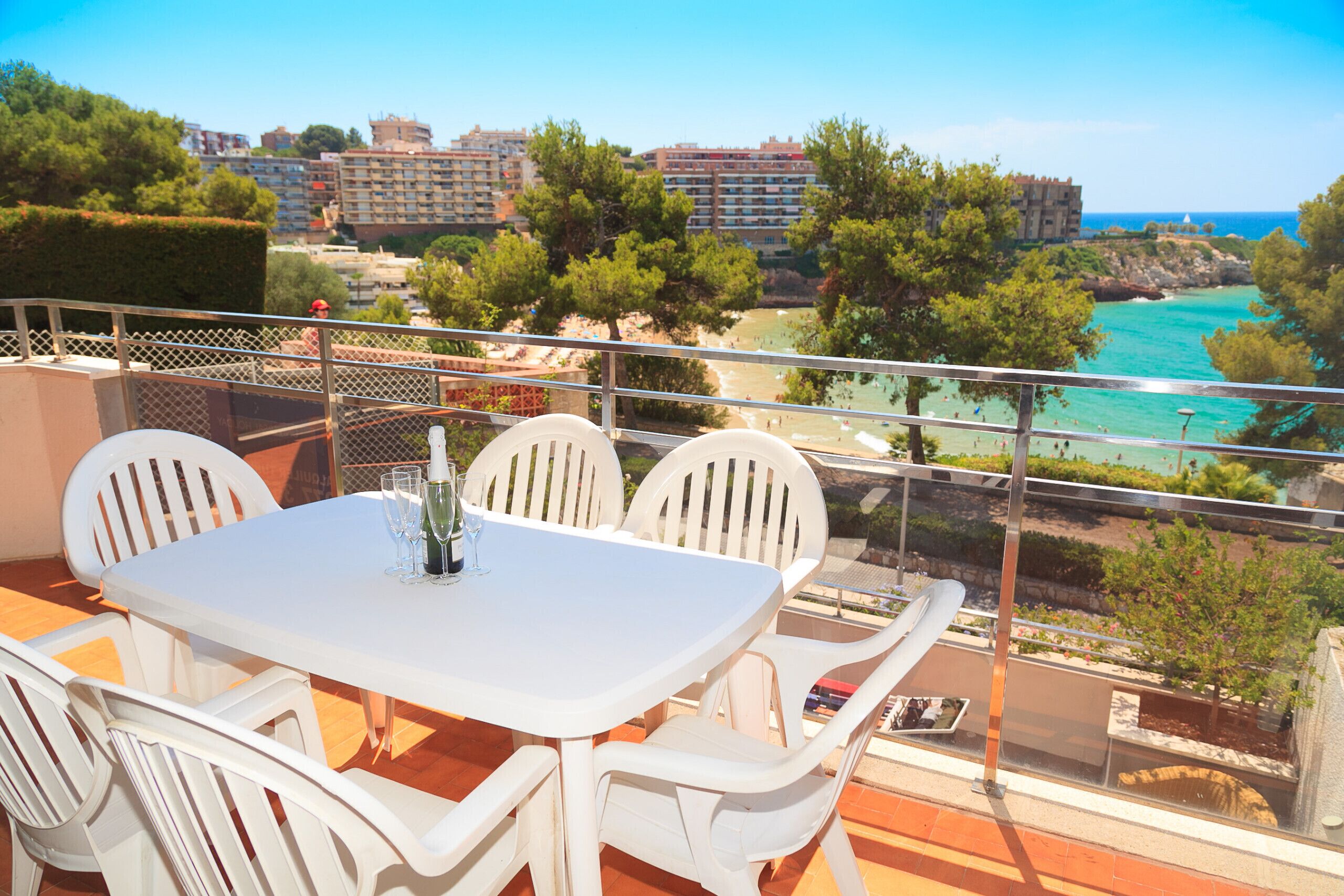 Un balcón con una mesa y sillas blancas, con vista a una playa y edificios.