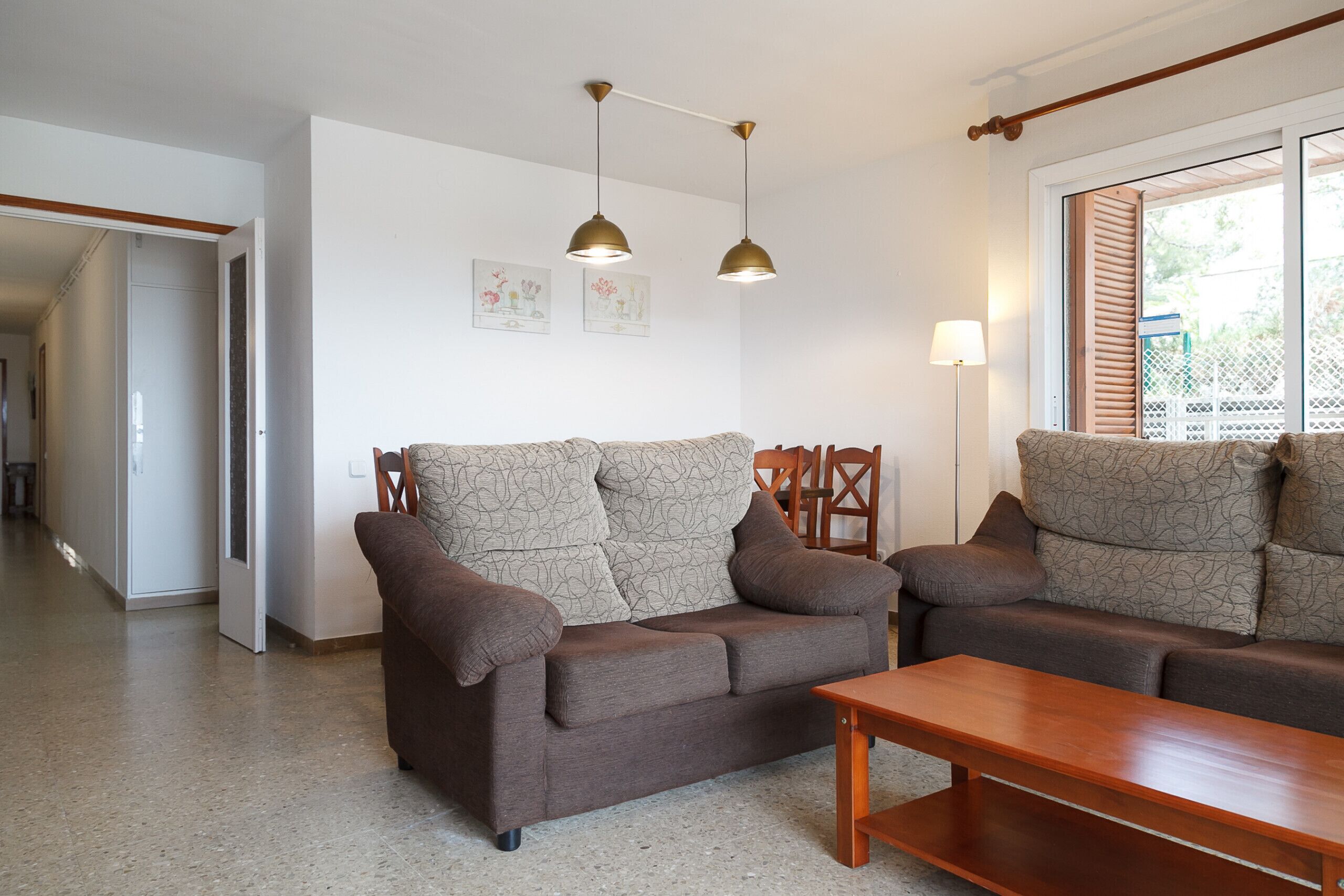 Una sala de estar con un sofá marrón, una mesa de centro de madera y dos lámparas colgantes. Hay una puerta que da a otra habitación y una ventana con persianas.
